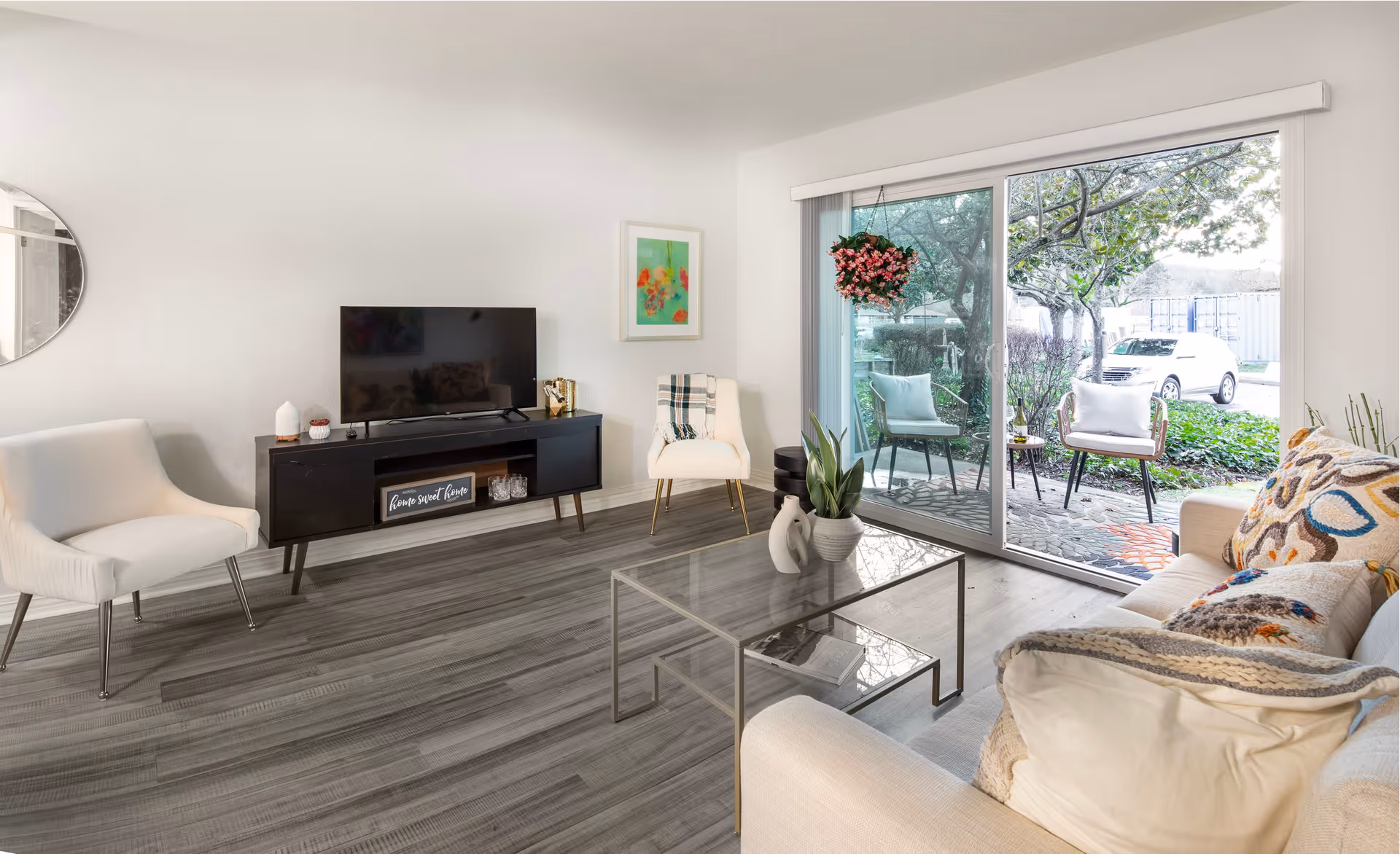 A bright and modern living room with a light-colored sofa adorned with patterned pillows and a throw blanket. There is a glass coffee table with decorative items on it. Two white chairs are positioned near a black TV stand holding a flat-screen television. A framed artwork hangs on the wall, and large sliding glass doors open to an outdoor patio area with chairs and greenery visible outside.