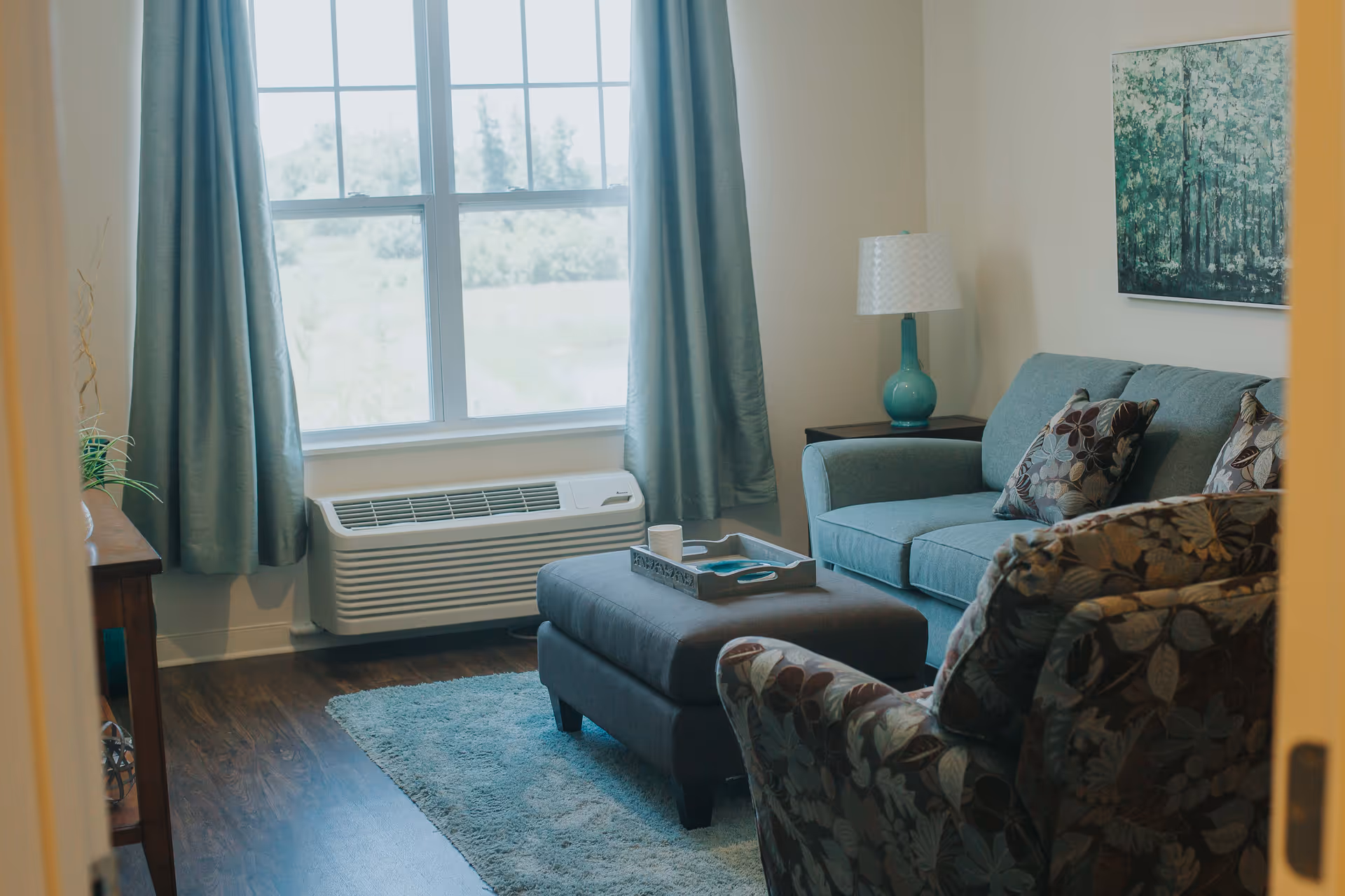 A cozy living room with a teal sofa adorned with floral patterned pillows, a matching floral armchair, a dark ottoman with a tray on top, a side table with a teal lamp, a large window with teal curtains, and a painting of a forest on the wall.