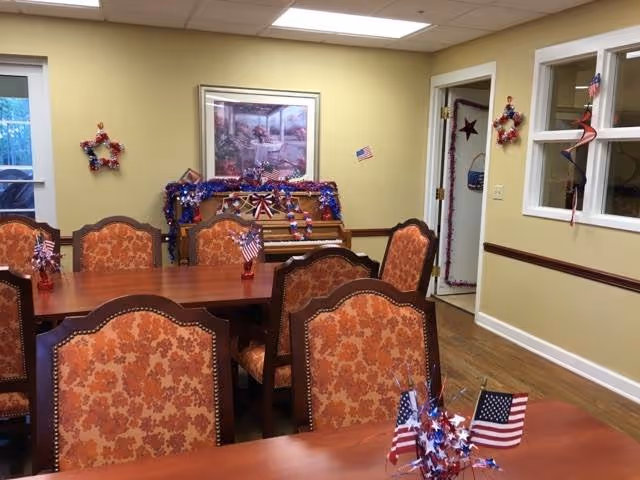 A dining room with wooden tables and upholstered chairs featuring an orange floral pattern. The room is decorated with patriotic American flags and red, white, and blue garlands. A piano against the wall is also adorned with similar decorations. The walls are painted light yellow with white trim, and there are windows and a door visible in the background.