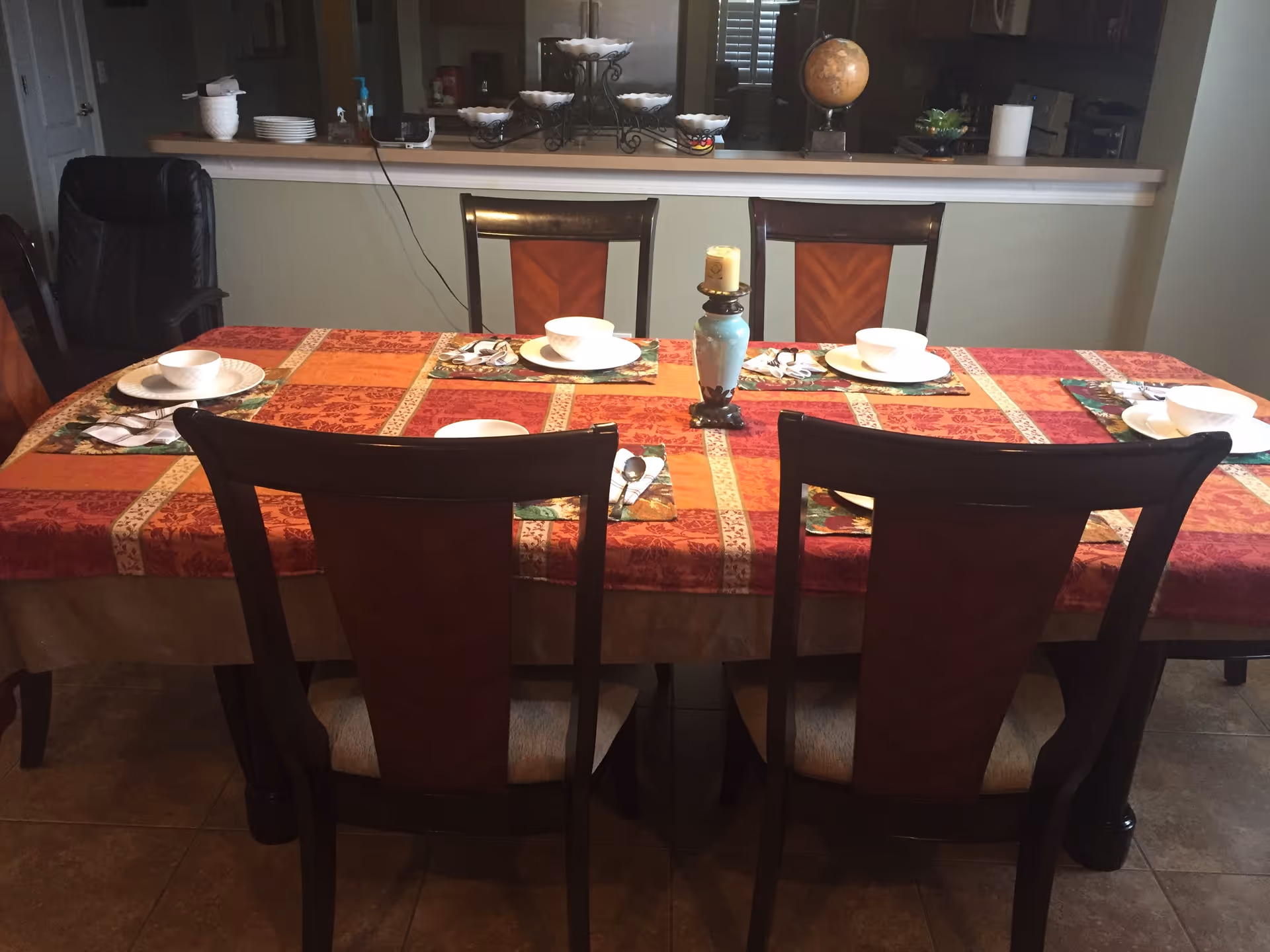Dining table set with a red and orange patterned tablecloth, six place settings with white bowls and plates, silverware on floral napkins, and a candle holder with a candle in the center. Behind the table is a counter with decorative items including a globe and bowls.