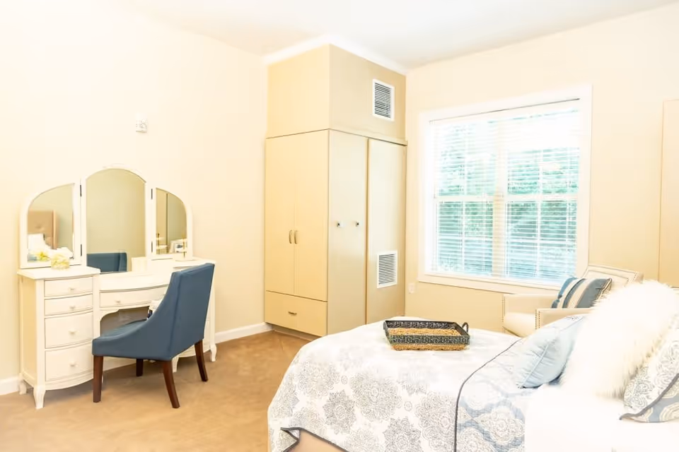 Bright bedroom with a bed and patterned bedding, a vanity with mirror and blue chair, a built-in wardrobe, and a window with blinds.