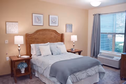 A cozy bedroom with a wooden bed frame, light blue bedding, and three decorative pillows. There are two wooden nightstands on either side of the bed, each with a lamp. The walls are beige with three framed pictures above the bed. A window with gray curtains and a white air conditioning unit is on the right side of the room.
