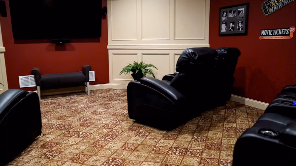 A cozy home theater room with patterned tile flooring, dark leather recliner chairs, a wall-mounted flat screen TV, a small bench against a red wall, a potted plant in the corner, and movie-themed wall decorations including framed black and white photos and signs that say 'CINEMA' and 'MOVIE TICKETS'.