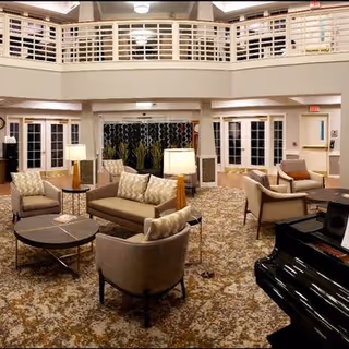 Spacious and well-lit living room area with multiple cushioned chairs and sofas arranged around small tables. A black grand piano is visible in the foreground on the right. The room features a patterned carpet, white walls, and a balcony railing on the upper level. Several lamps provide warm lighting, and large windows and doors are seen in the background.