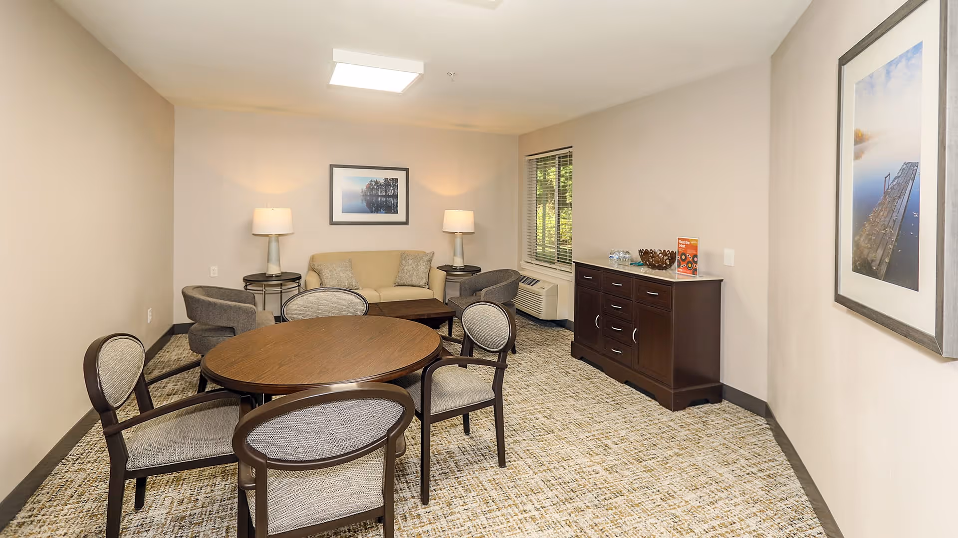 A cozy living room area with a round wooden table surrounded by four upholstered chairs in the foreground. In the background, there is a beige sofa with two cushions, two side tables each with a lamp, and two gray armchairs. A dark wooden cabinet with drawers is placed against the right wall under a window with blinds. The walls are decorated with framed landscape photographs, and the floor is carpeted with a patterned design.