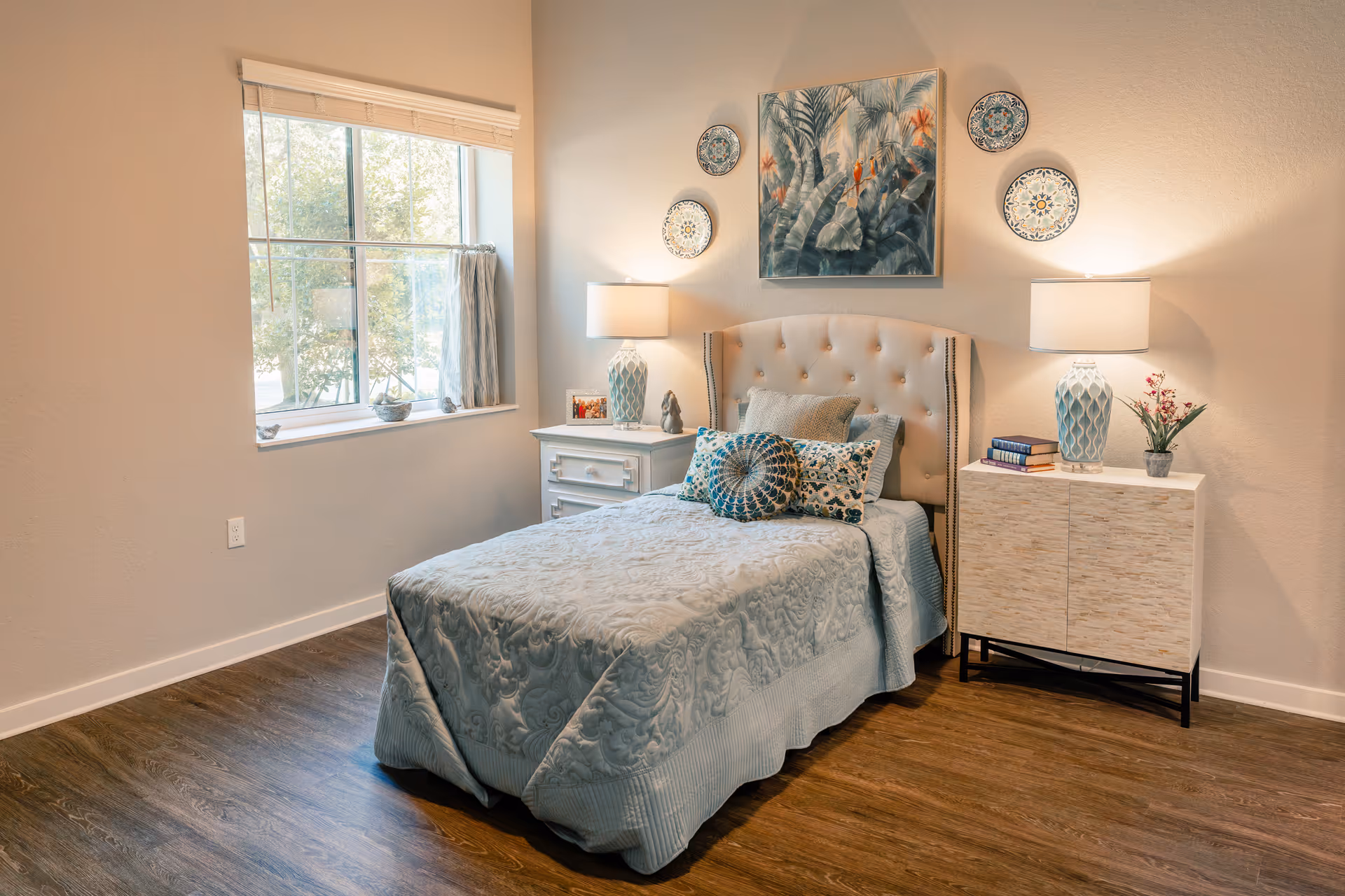 A cozy bedroom with a single bed covered in a light blue quilt and decorative pillows. The bed has a tufted headboard and is flanked by two nightstands, each with a lamp. Above the bed, there is a painting of tropical birds and plants, surrounded by decorative plates on the wall. A window with blinds and a curtain lets in natural light, and the room has wooden flooring.