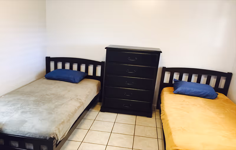 A small bedroom with two single beds placed against adjacent walls. Each bed has a dark wooden frame and a blue pillow. Between the beds is a black chest of drawers with five drawers. The floor is tiled with light-colored tiles, and the walls are plain white.