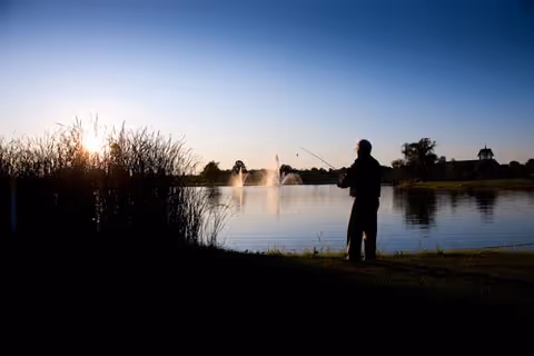 Silhouette of a person fishing by a lake at sunset with water fountains and reeds in the background.