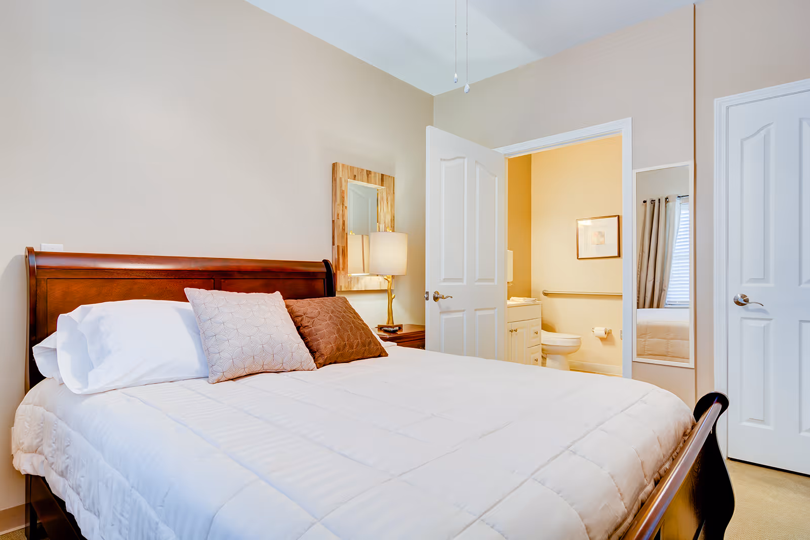 A bedroom with a wooden bed frame, white bedding, and two decorative pillows. A bedside table with a lamp and a wooden framed mirror is next to the bed. The door to an adjoining bathroom is open, showing a toilet, vanity, and a framed picture on the wall. Another closed door and a full-length mirror are visible on the right side of the room.