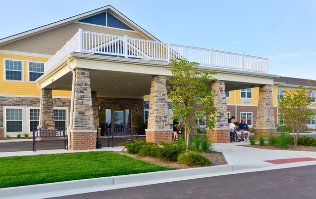 Exterior view of a senior living facility named Traditions at Reagan Park, showing a covered entrance supported by stone pillars, a well-maintained lawn, and a few people sitting and standing near the entrance.
