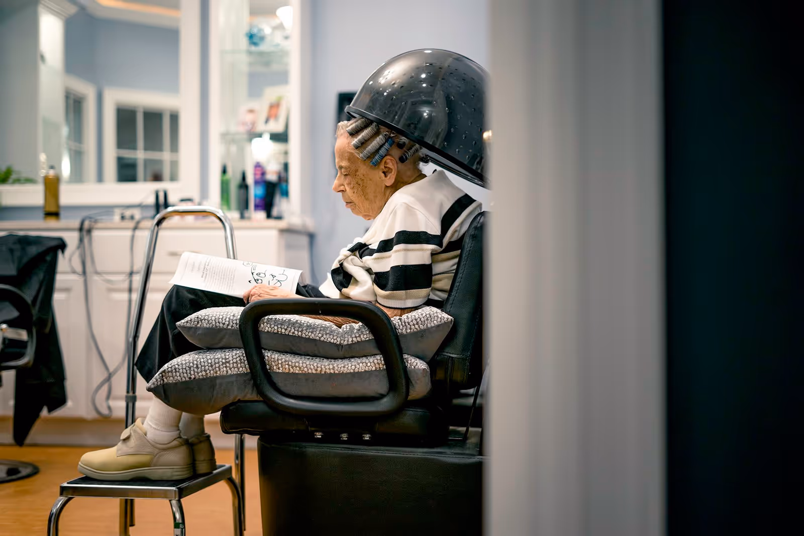 An elderly woman sitting under a hair dryer in a salon chair, reading a book. She has hair rollers in her hair and is resting her feet on a small stool. The background shows salon equipment and shelves with hair products.