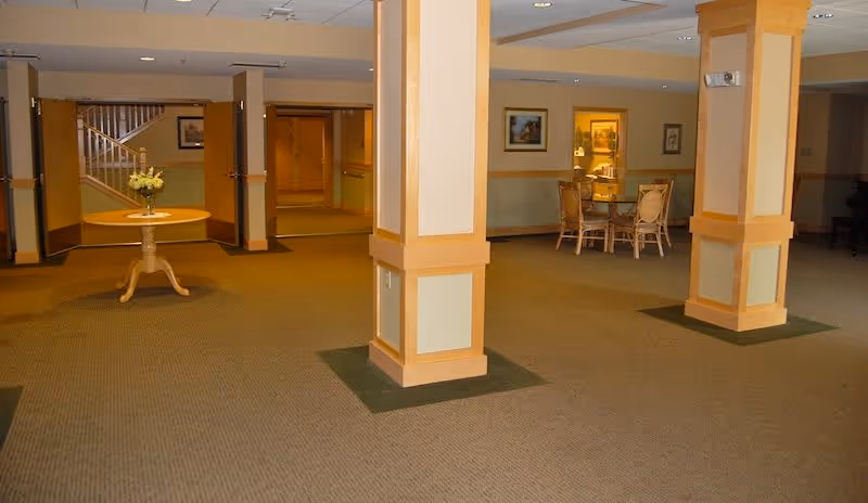 Spacious interior common area with beige carpet and cream-colored walls accented with wooden trim. Two large square columns are in the foreground. In the background, there is a round table with a vase of flowers and a seating area with a table and chairs. A staircase and framed artwork are visible on the walls.