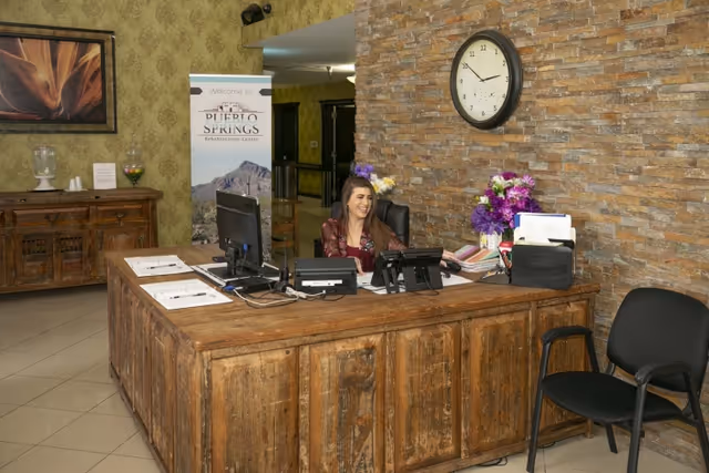 Reception area of Pueblo Springs Rehabilitation Center with a wooden front desk, a smiling receptionist seated behind the desk, a wall clock showing 10:10, a bouquet of purple flowers, office phones, and a banner with the facility's name in the background.