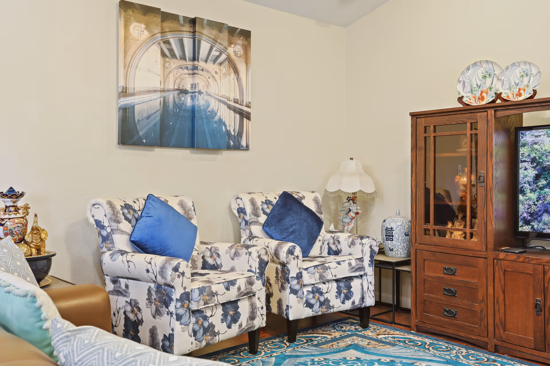 A cozy living room area with two floral-patterned armchairs each with a blue cushion, a wooden cabinet with decorative plates on top, a table lamp with a lace shade, and a wall art piece depicting an indoor swimming pool.