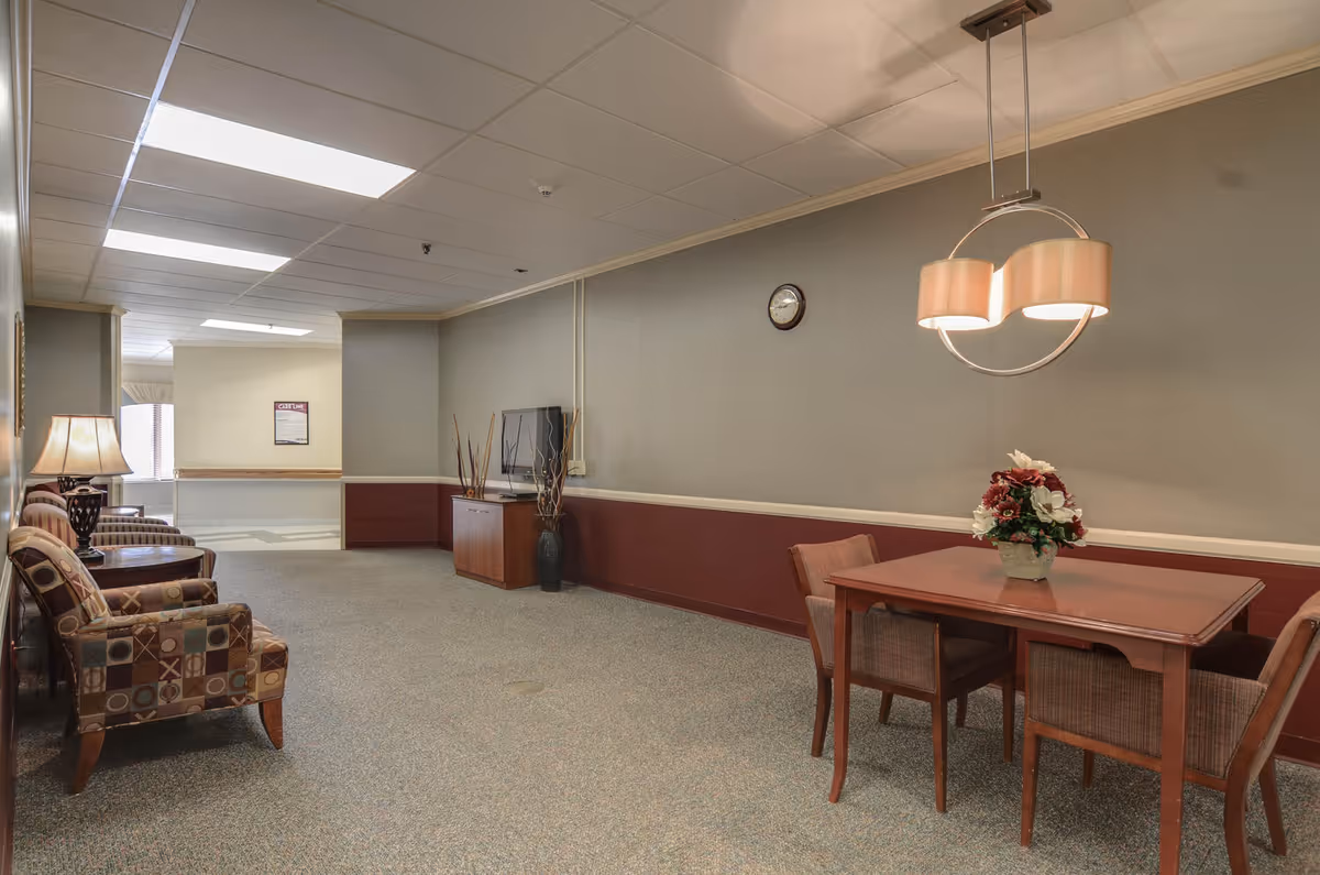 A quiet common area in Beckley Healthcare Center featuring a small wooden table with four chairs and a floral centerpiece. On the left side, there are three patterned armchairs with a lamp on a side table. A flat-screen TV is mounted on the wall above a wooden cabinet, and a clock hangs on the wall. The room has a beige and maroon color scheme with carpeted flooring and fluorescent ceiling lights.