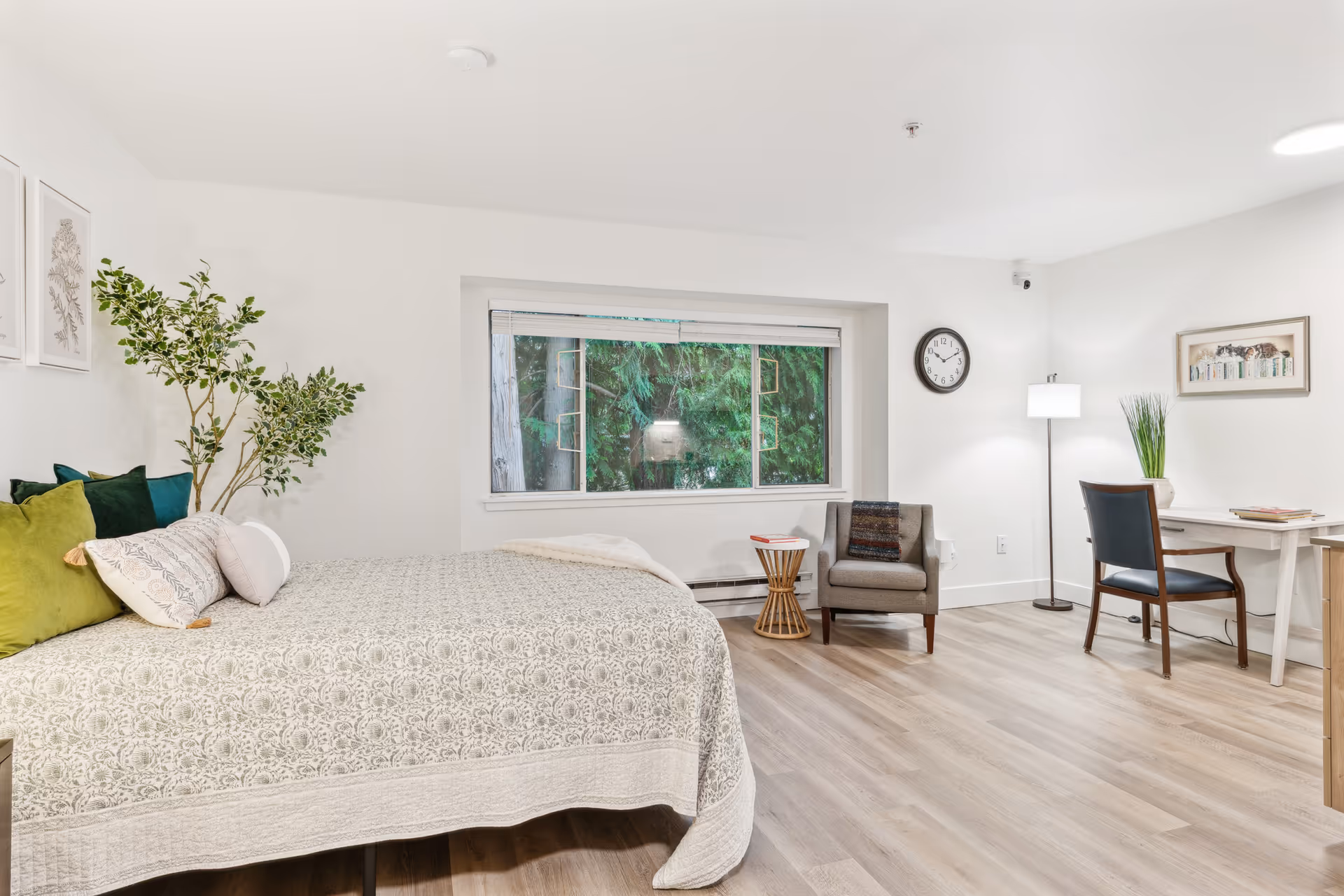 A bright and spacious bedroom with a large bed covered in a patterned beige quilt and multiple pillows in green, white, and blue. There is a potted plant next to the bed and two framed botanical prints on the wall. A large window shows green trees outside. The room also features a gray armchair with a small round wooden side table, a floor lamp, a wall clock, and a desk with a chair and decorative items on it. The floor is light wood and the walls are white.
