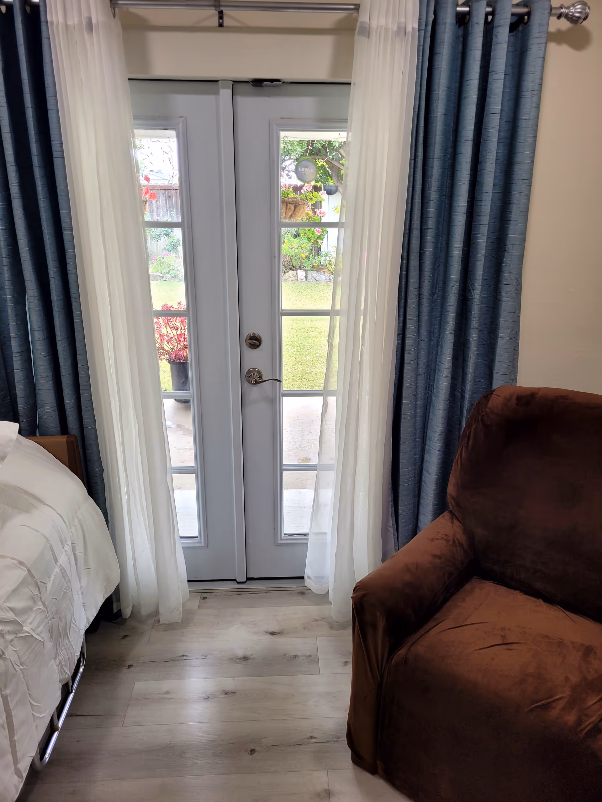 A cozy room with a brown armchair on the right and a bed with white bedding on the left. In the center, there are white French doors with glass panes, framed by sheer white curtains and dark blue drapes. Outside the doors, a garden with green grass and hanging flower baskets is visible.