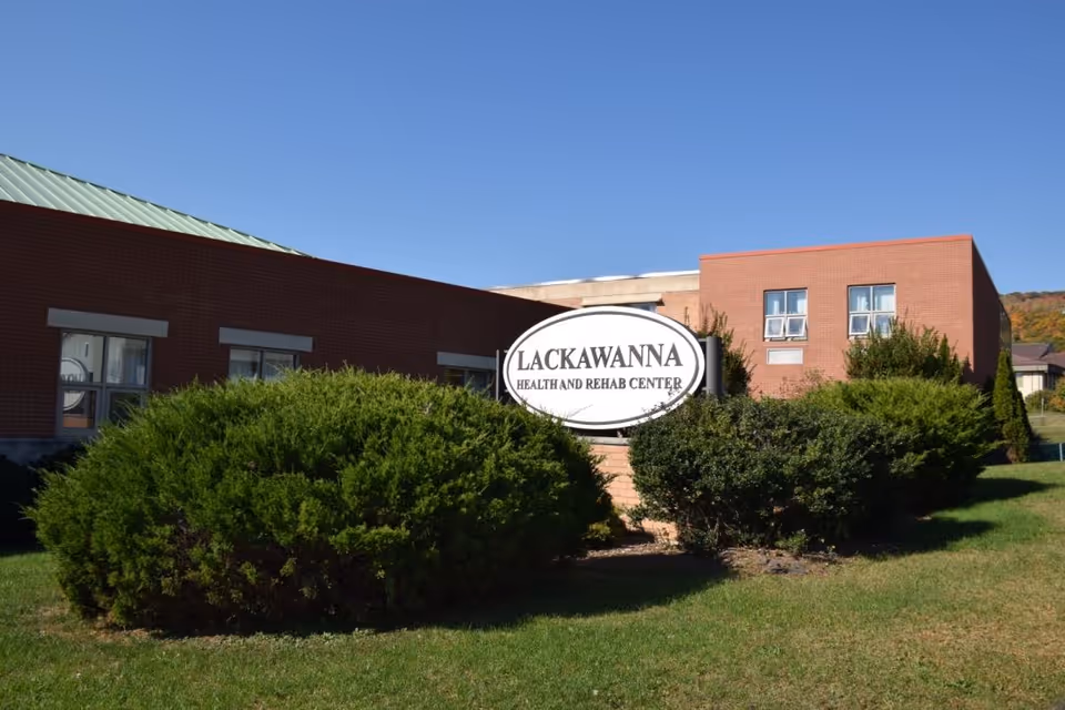 Exterior view of the Lackawanna Health and Rehab Center building with a clear blue sky. The building is made of red brick with several windows, and there are green bushes and grass in the foreground. A white oval sign with black text reading 'Lackawanna Health and Rehab Center' is prominently displayed among the bushes.