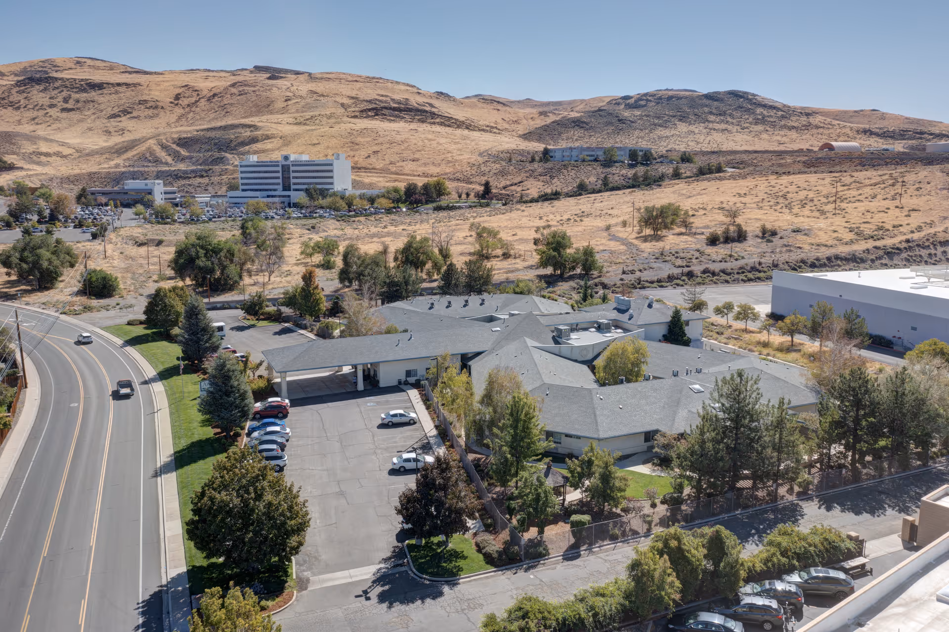 Aerial view of Arbors Memory Care facility surrounded by trees and parking lots, with a road curving along the left side and dry hills in the background under a clear blue sky.