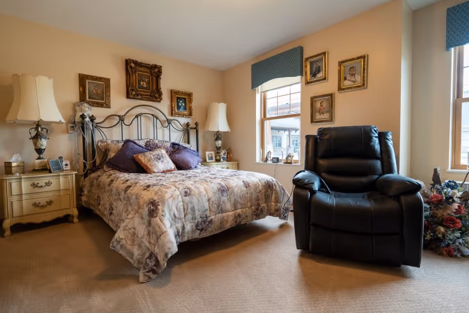 A cozy bedroom with a floral-patterned bedspread on a metal bed frame, two bedside tables with ornate lamps, framed pictures on the walls, a large black recliner chair, and two windows with blue valances letting in natural light.