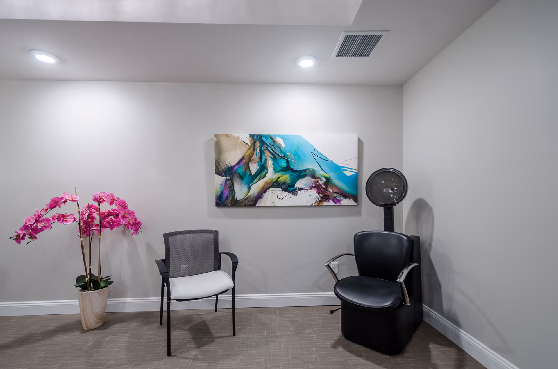 Small seating area with two chairs, a standing hair dryer, a colorful abstract painting, and a potted pink orchid against a neutral wall.