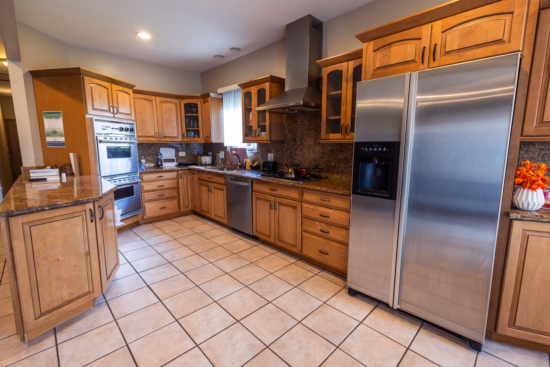 A spacious kitchen with wooden cabinets, granite countertops, tiled floor, stainless steel double-door refrigerator, built-in oven, dishwasher, and a range hood above the stove. There is a window with vertical blinds letting in natural light and a vase with orange flowers on the counter.