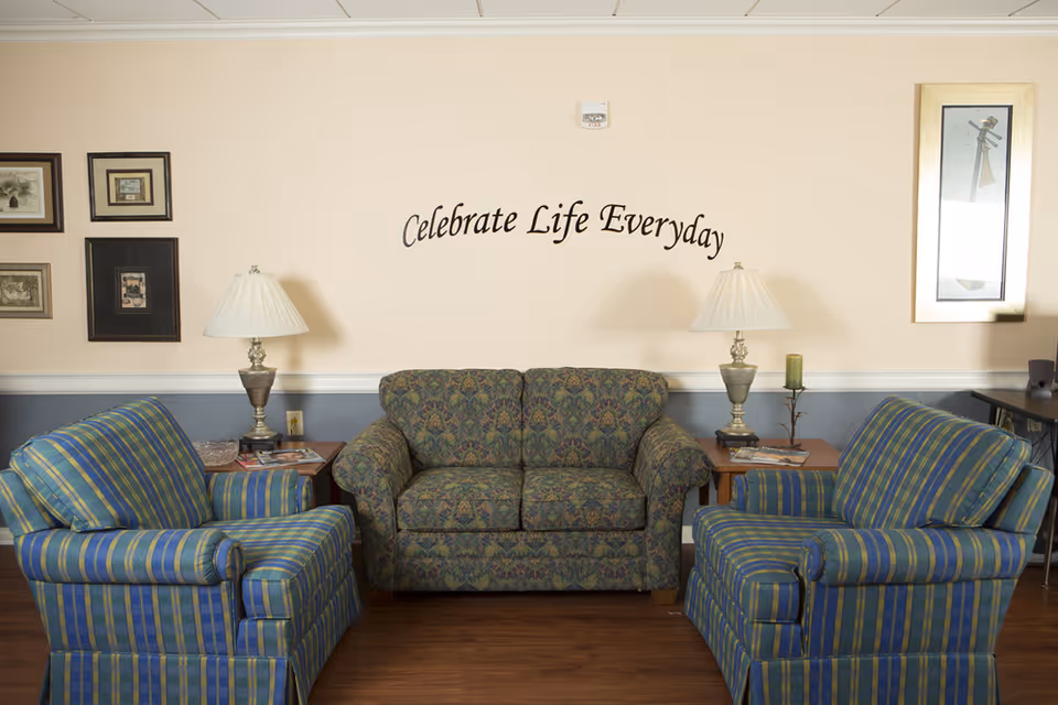 A cozy sitting area with a patterned loveseat flanked by two blue and green striped armchairs. Two side tables each hold a lamp with a white shade. The wall behind the loveseat has framed artwork and a decorative sign that reads 'Celebrate Life Everyday.'