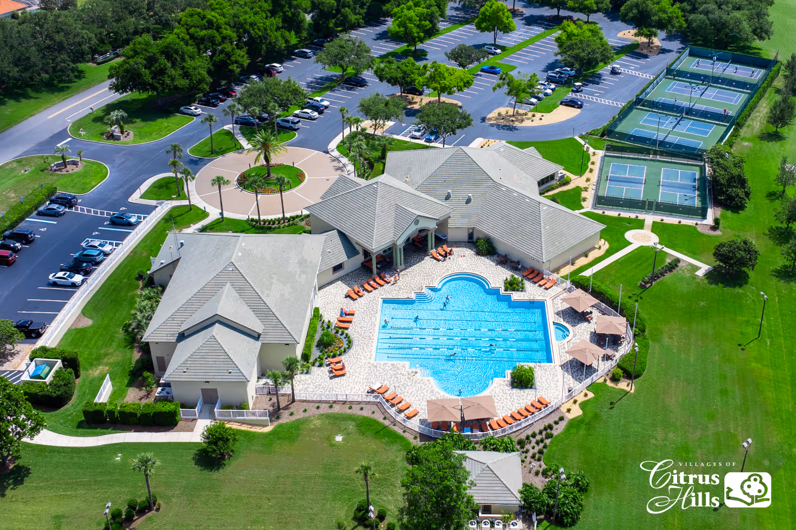 Aerial view of The Villages of Citrus Hills facility showing a large swimming pool with lounge chairs and umbrellas around it, adjacent buildings with gray roofs, multiple tennis and pickleball courts, parking lots with cars, and green landscaped areas with trees and pathways.