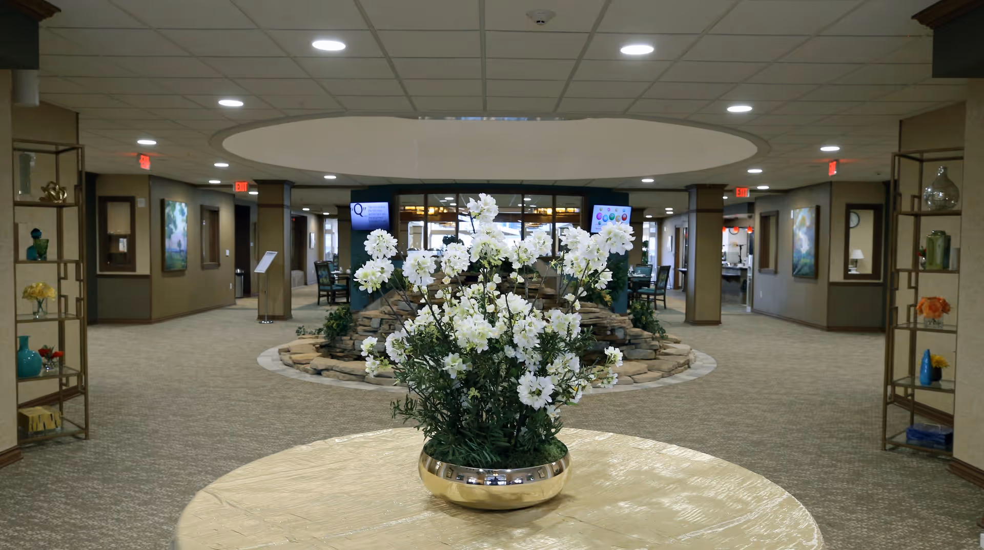 Interior view of a spacious common area in a retirement community with a round table in the foreground holding a large vase of white flowers. The room has beige carpeting, decorative shelving on both sides with various ornaments, and a circular stone feature in the center. Multiple doorways and hallways lead off the main area, and there are two digital screens mounted on a wall in the background.