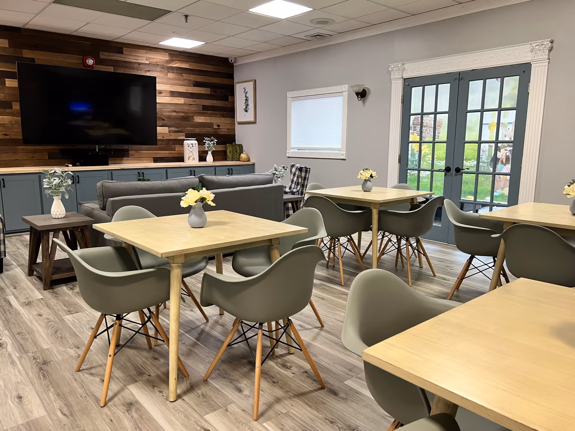 A bright and modern common area in an assisted living facility featuring several light wood tables with gray chairs around them. Each table has a small vase with yellow flowers. In the background, there is a gray couch facing a large flat-screen TV mounted on a wooden accent wall. French doors with glass panes lead to an outdoor garden area visible through the glass. The room has light wood flooring and soft gray walls.