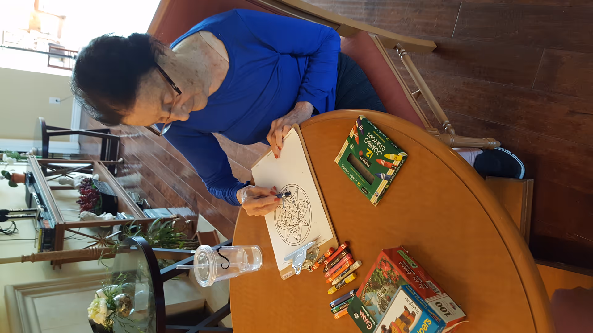 An older woman sitting at a round table coloring a mandala page with crayons in a communal living area.