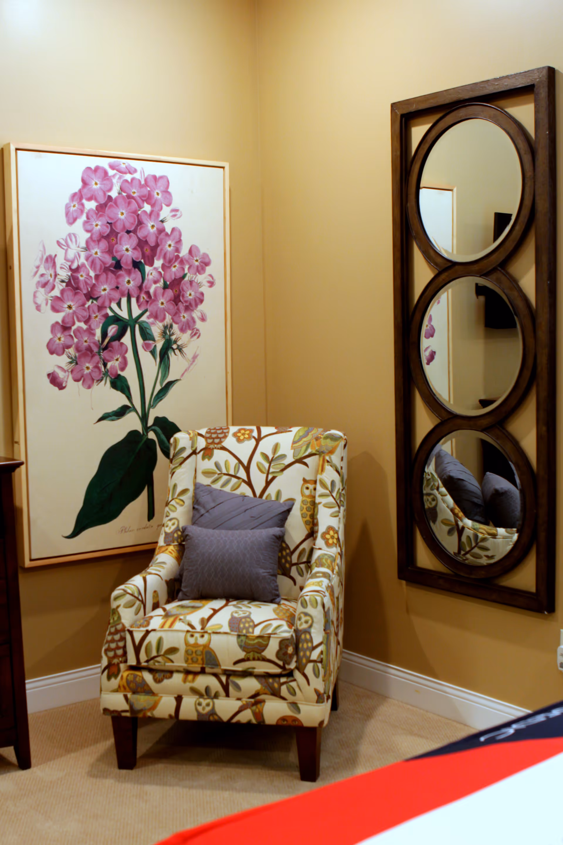 A cozy corner of a room featuring a patterned armchair with owl and branch designs, two gray cushions on the chair, a large framed artwork of pink flowers on the wall, and a vertical wooden-framed mirror with three circular sections reflecting parts of the chair and artwork.