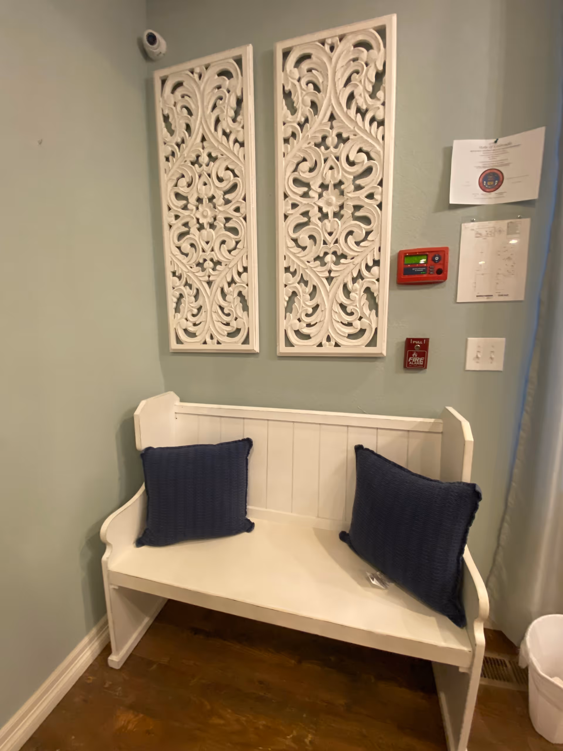 A small white wooden bench with two dark blue cushions placed on it, positioned against a light green wall. Above the bench are two decorative white carved wooden panels. To the right on the wall are a red fire alarm pull station, a red fire alarm control panel, and two documents pinned to the wall. The floor is wooden.
