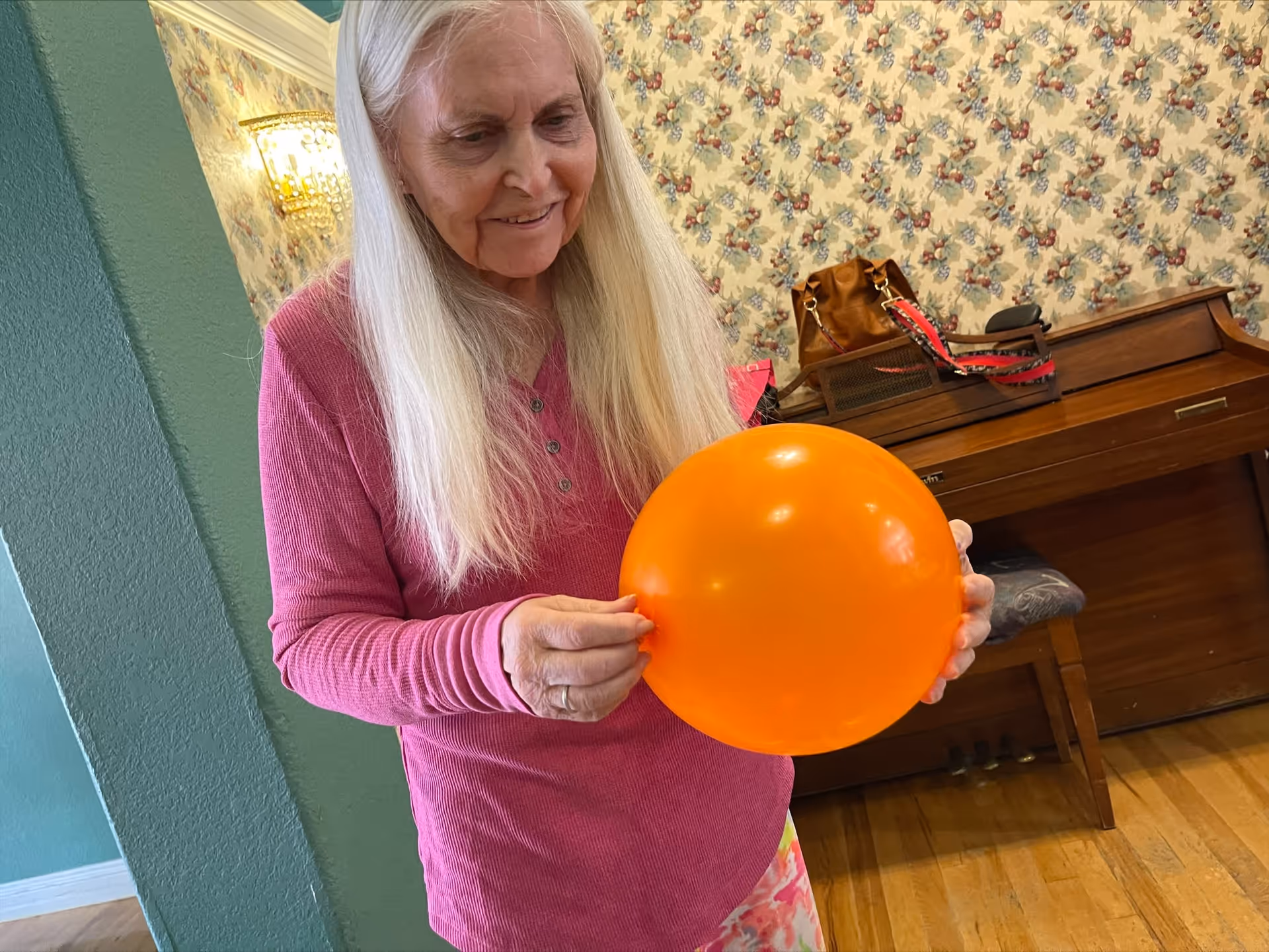 An elderly woman with long white hair wearing a pink long-sleeve shirt is holding an orange balloon indoors. Behind her is a wooden piano with a brown handbag and a red strap on top, and floral wallpaper on the wall.