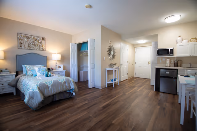 A spacious studio apartment with a single bed dressed in blue and beige bedding, a nightstand with a lamp, an open closet with shelves, a small table with decorative items, and a kitchenette area with a microwave, mini fridge, and white cabinets. The room has wood flooring and neutral-colored walls.