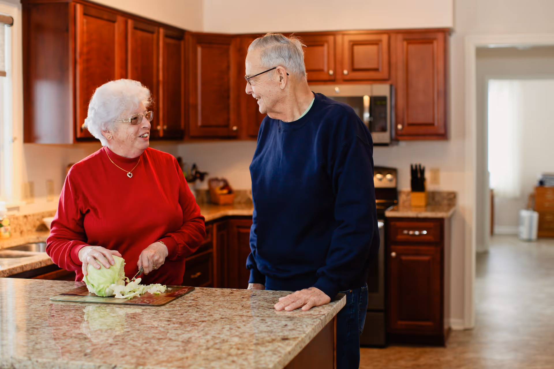 An elderly woman in a red sweater is chopping lettuce on a cutting board in a kitchen while talking to an elderly man in a navy blue sweater who is leaning on the kitchen island. The kitchen has wooden cabinets, granite countertops, and stainless steel appliances.