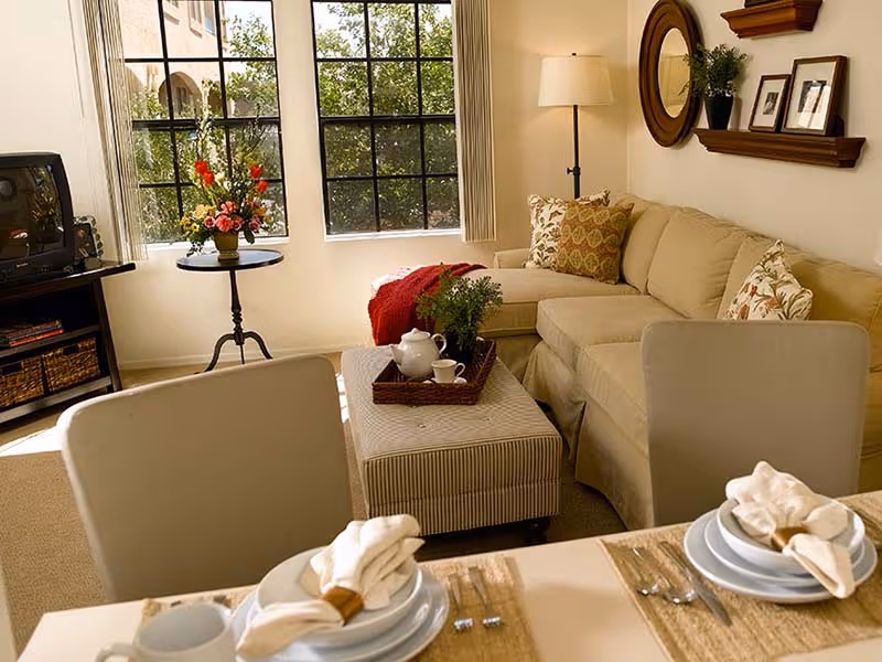 Cozy living room with a beige sectional sofa, ottoman coffee table, dining place settings in the foreground, and a large window letting in daylight.