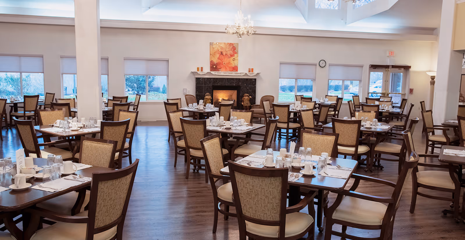 A spacious dining room in a senior living community with multiple wooden tables and chairs arranged neatly. Each table is set with white cups, glasses, and silverware. The room features large windows with blinds, a fireplace with decorative items and a floral painting above it, and a chandelier hanging from the ceiling. The floor is wooden, and the room is well-lit with natural light.