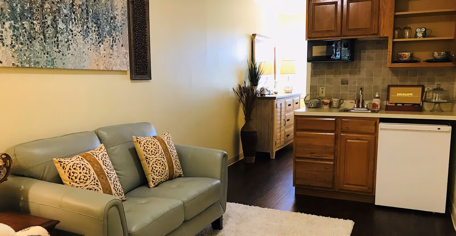A cozy living area with a light green leather loveseat adorned with two decorative pillows. To the right, there is a small kitchen area featuring wooden cabinets, a white mini refrigerator, a microwave, and a sink. The floor is dark wood, and a white rug is partially visible. In the background, a wooden dresser with a mirror and two lamps is seen, along with a tall vase holding decorative dried plants. A large abstract painting hangs on the wall above the loveseat.