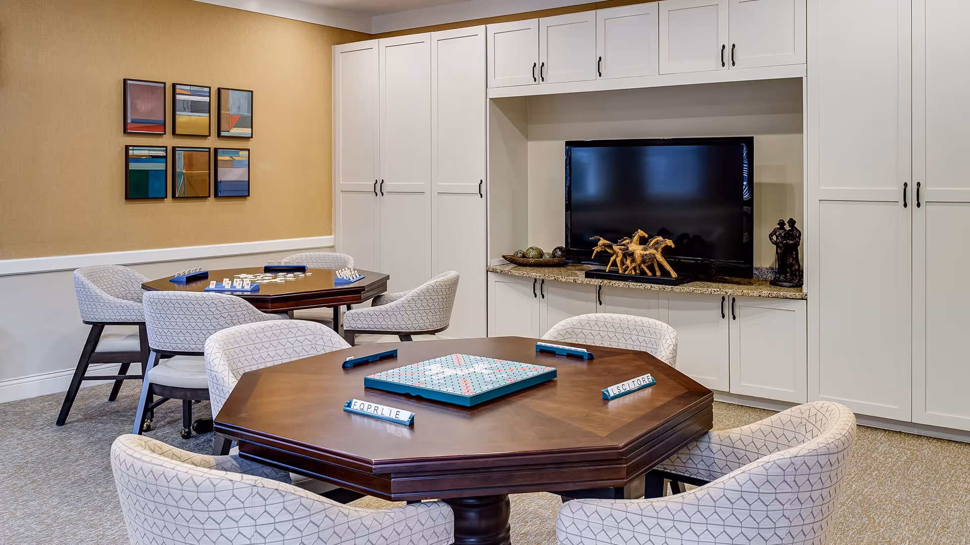A cozy game room with two wooden tables set up for board games, surrounded by cushioned chairs with patterned upholstery. The room features built-in white cabinets and a flat-screen TV on a granite countertop. Decorative items and colorful abstract artwork adorn the walls.