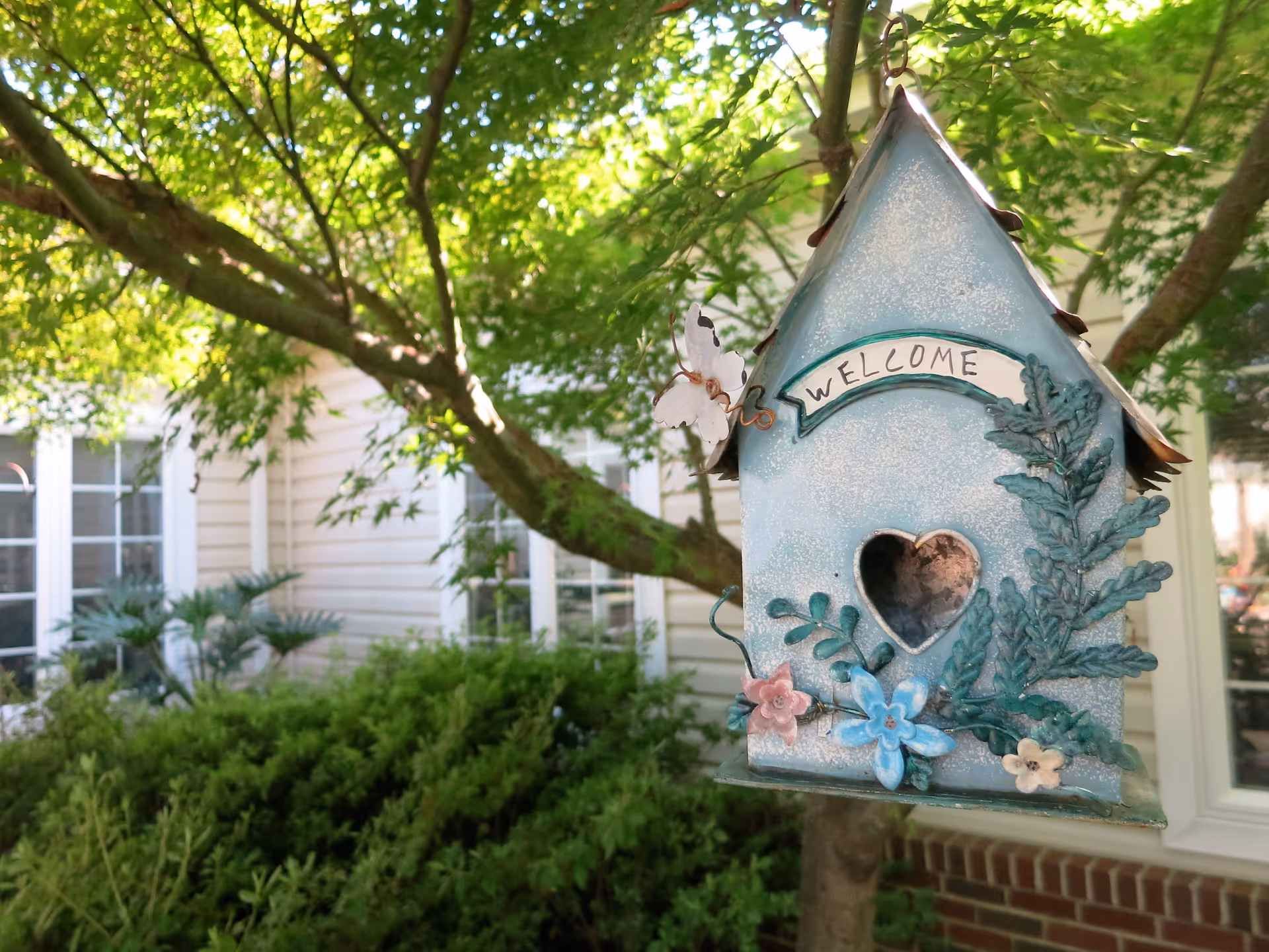 A decorative blue birdhouse with a heart-shaped hole and 'WELCOME' hangs from a tree branch in a green courtyard outside a building.