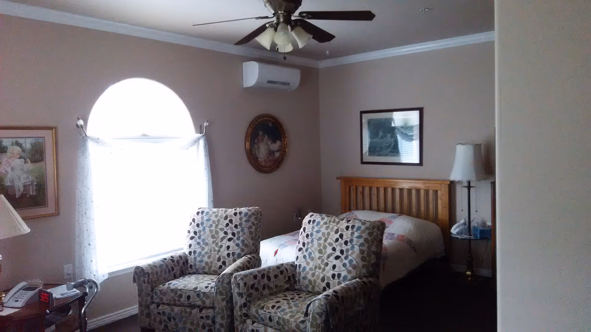 Furnished bedroom with a twin bed, two patterned armchairs by a large arched window, a ceiling fan, and bedside lamp.
