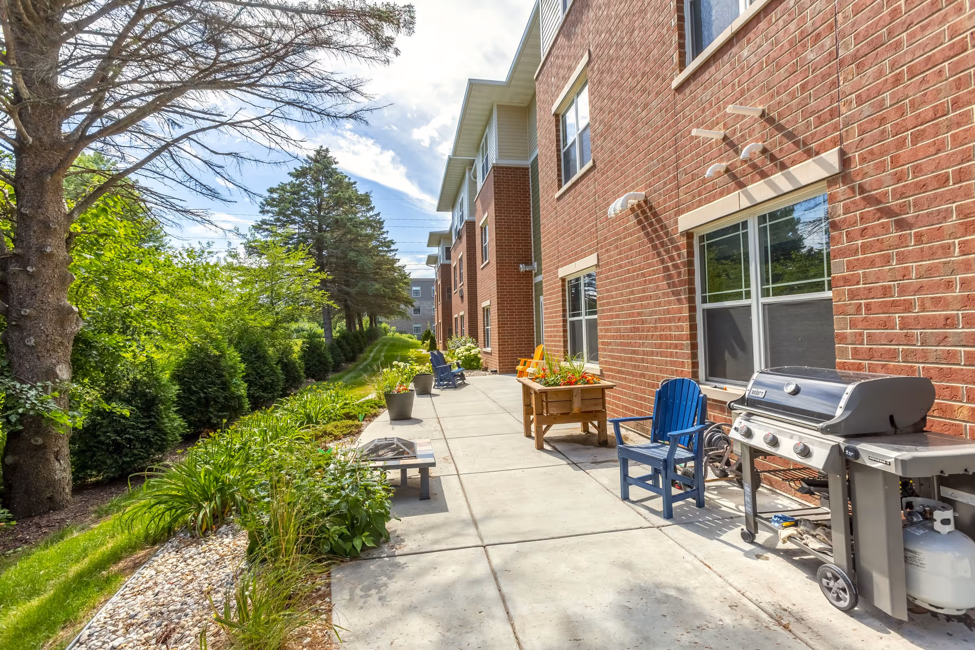 Outdoor patio area alongside a brick building with several colorful chairs, a grill, potted plants, and greenery including trees and shrubs under a partly cloudy sky.