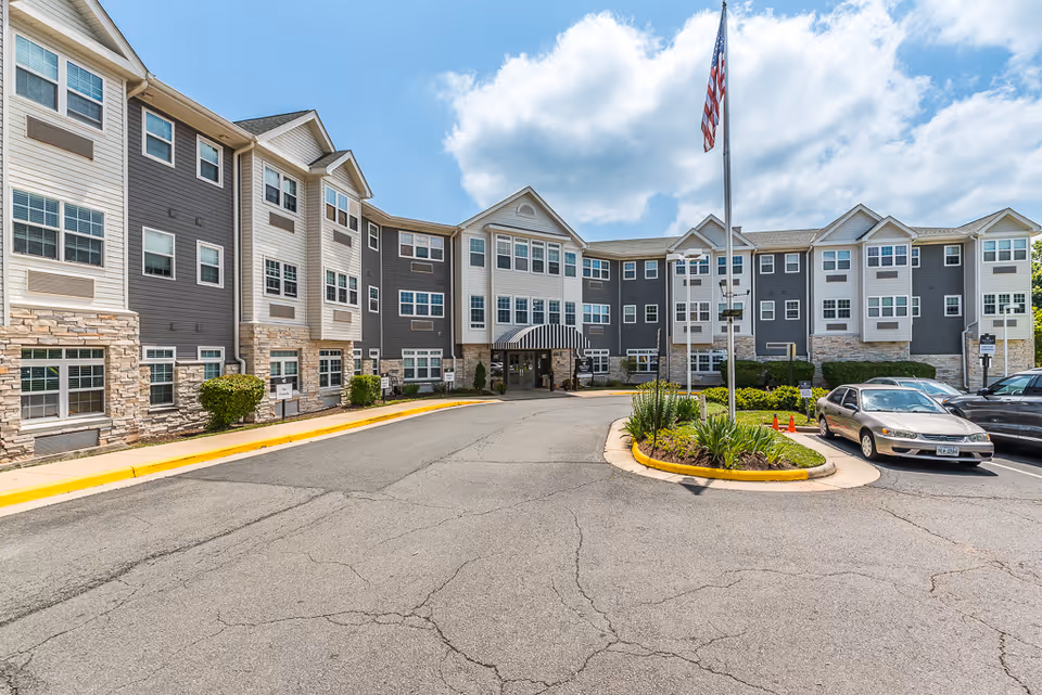 Exterior view of a three-story senior living facility building with a driveway and parking area in front. The building has a combination of gray and white siding with stone accents on the lower part. There is a flagpole with an American flag in the center of a landscaped roundabout near the entrance. Several cars are parked on the right side under a partly cloudy sky.