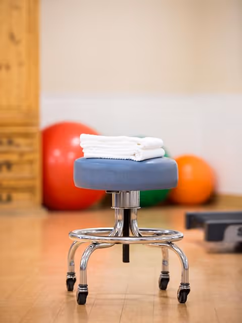 A rolling stool topped with folded white towels in a fitness/therapy room with colorful exercise balls in the background.