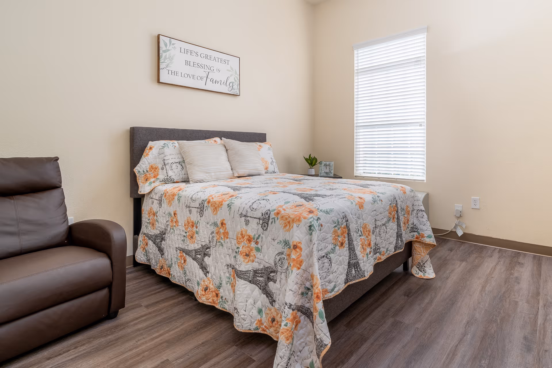 A bright bedroom with a double bed covered in a floral quilt, a brown recliner, wall art, and a window with blinds.
