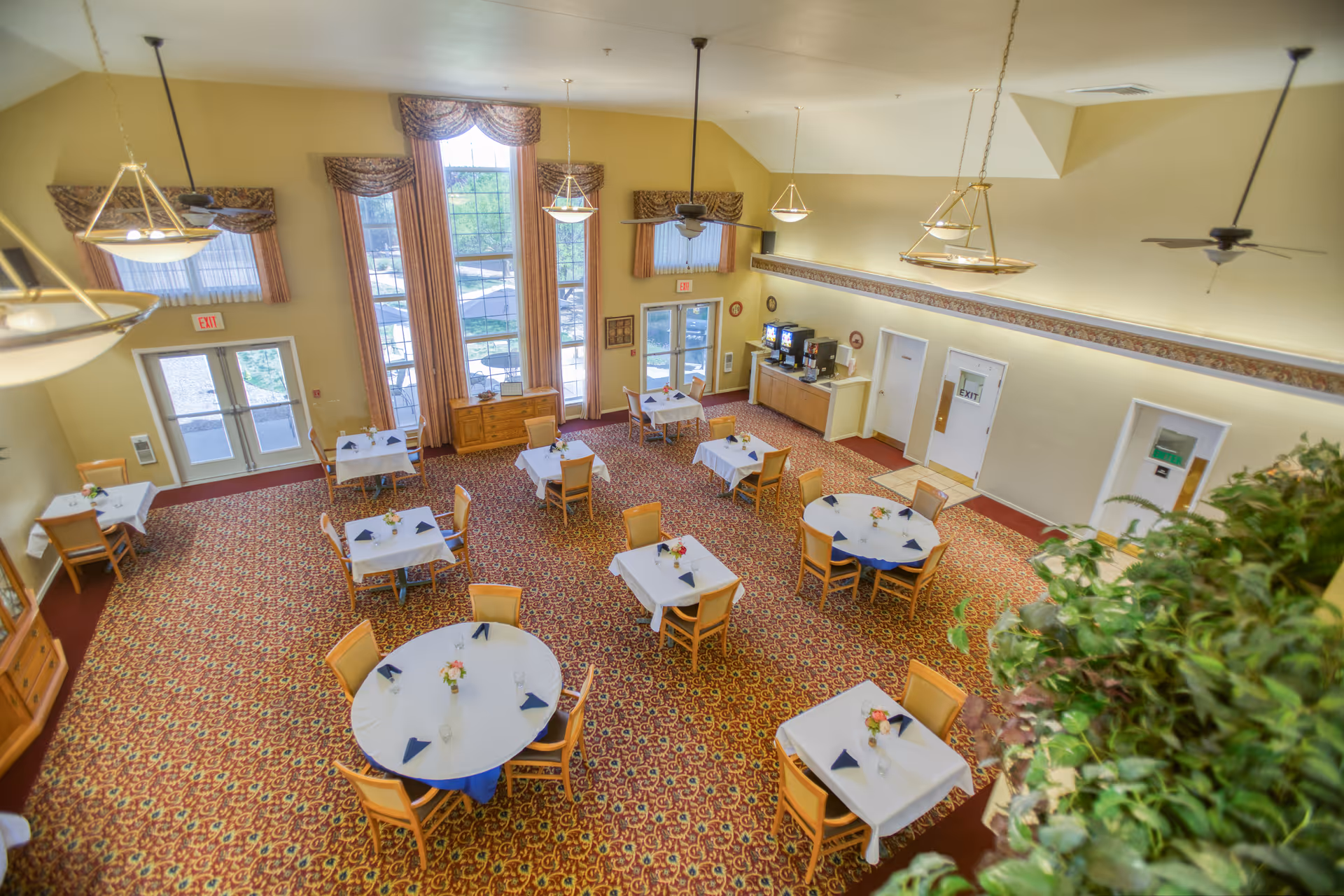 A spacious dining room with multiple tables covered in white tablecloths and set with napkins and small floral centerpieces. The room features large windows with drapes, patterned carpet, ceiling fans, and hanging light fixtures. There is a beverage station along one wall and several exit doors.