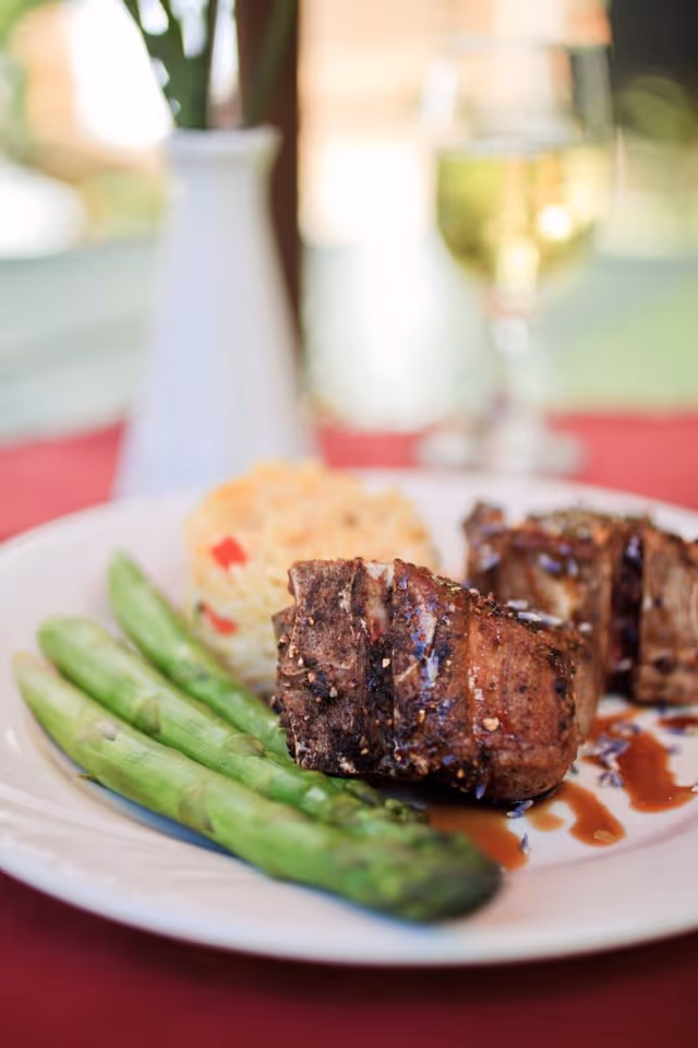 Close-up of a plate with grilled steak pieces, three asparagus spears, and a serving of rice with red bell peppers, with a blurred background featuring a white vase and a glass of white wine.