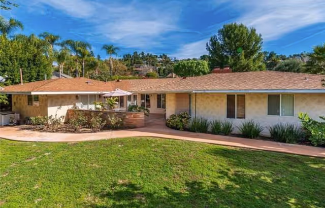 Single-story building with a tan exterior and brown shingle roof, surrounded by green grass and landscaping with shrubs and small trees under a blue sky with some clouds.