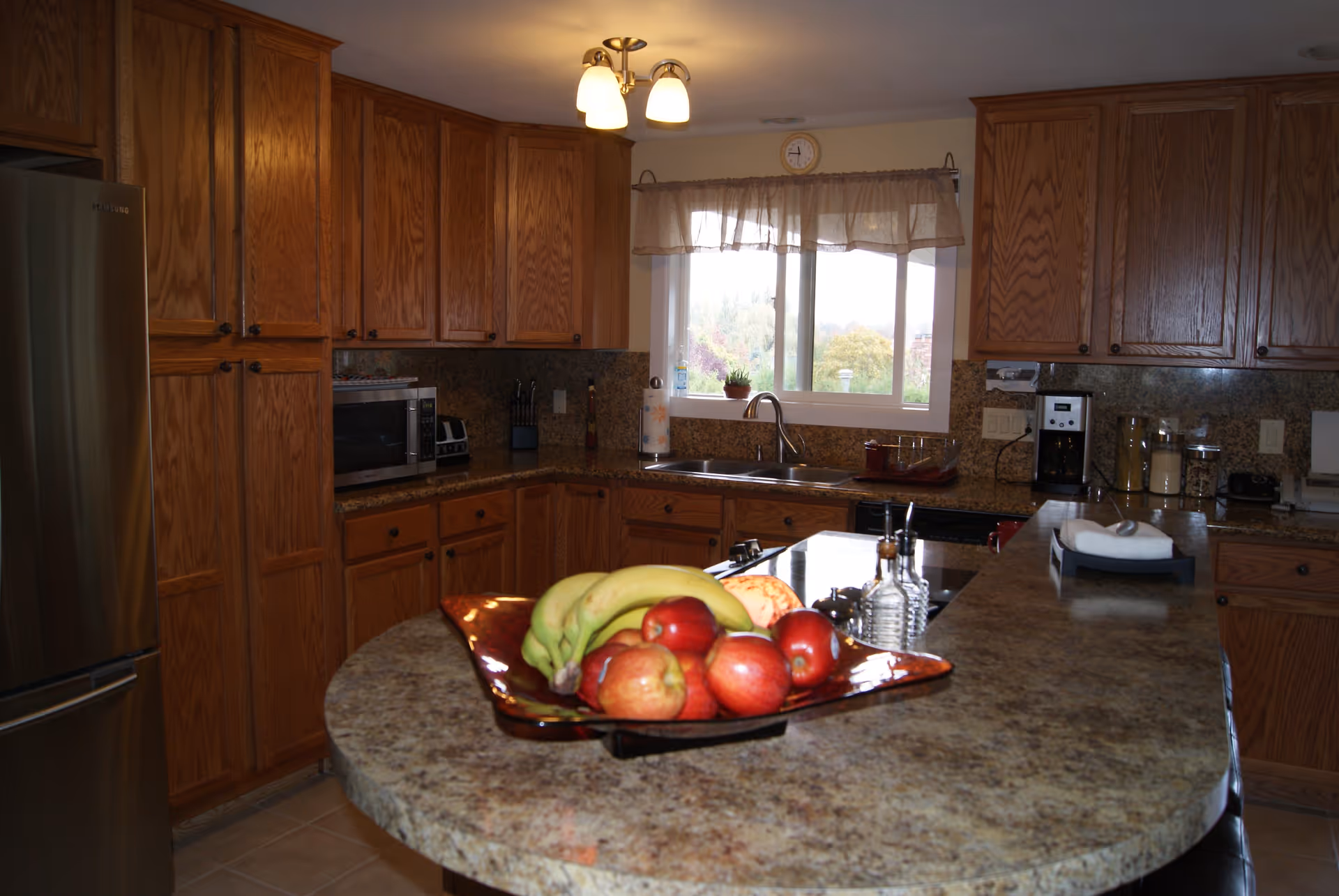 A kitchen with wooden cabinets, a granite countertop island with a fruit bowl containing bananas and apples, a stainless steel refrigerator, a microwave, a coffee maker, and a window with a sheer curtain letting in natural light.